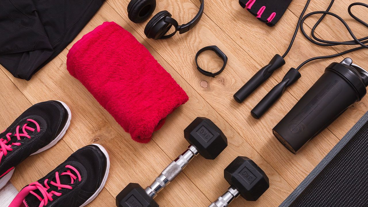 Fitness gear on wooden floor: sneakers, dumbbells, jump rope, headphones, towel, gloves, shaker bottle, and fitness tracker.