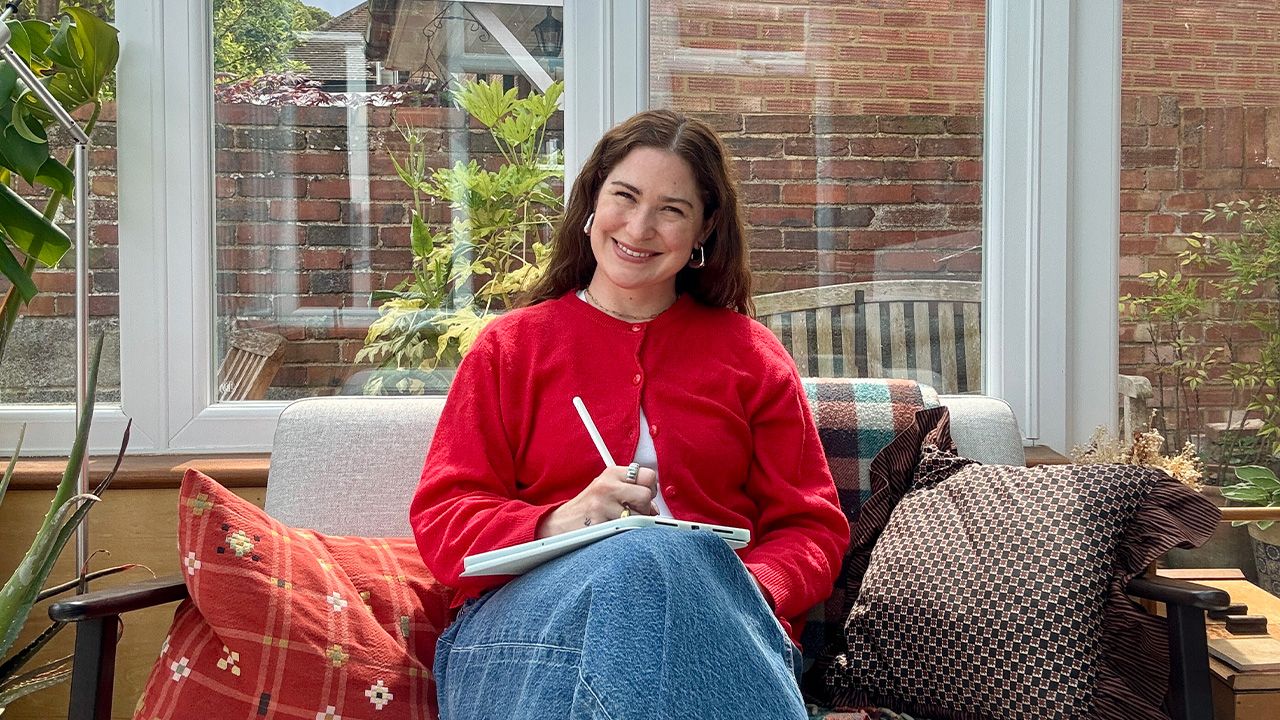 A woman in a red sweater sits on a sofa in a sunlit room, smiling and writing on a tablet. She is surrounded by colorful cushions.