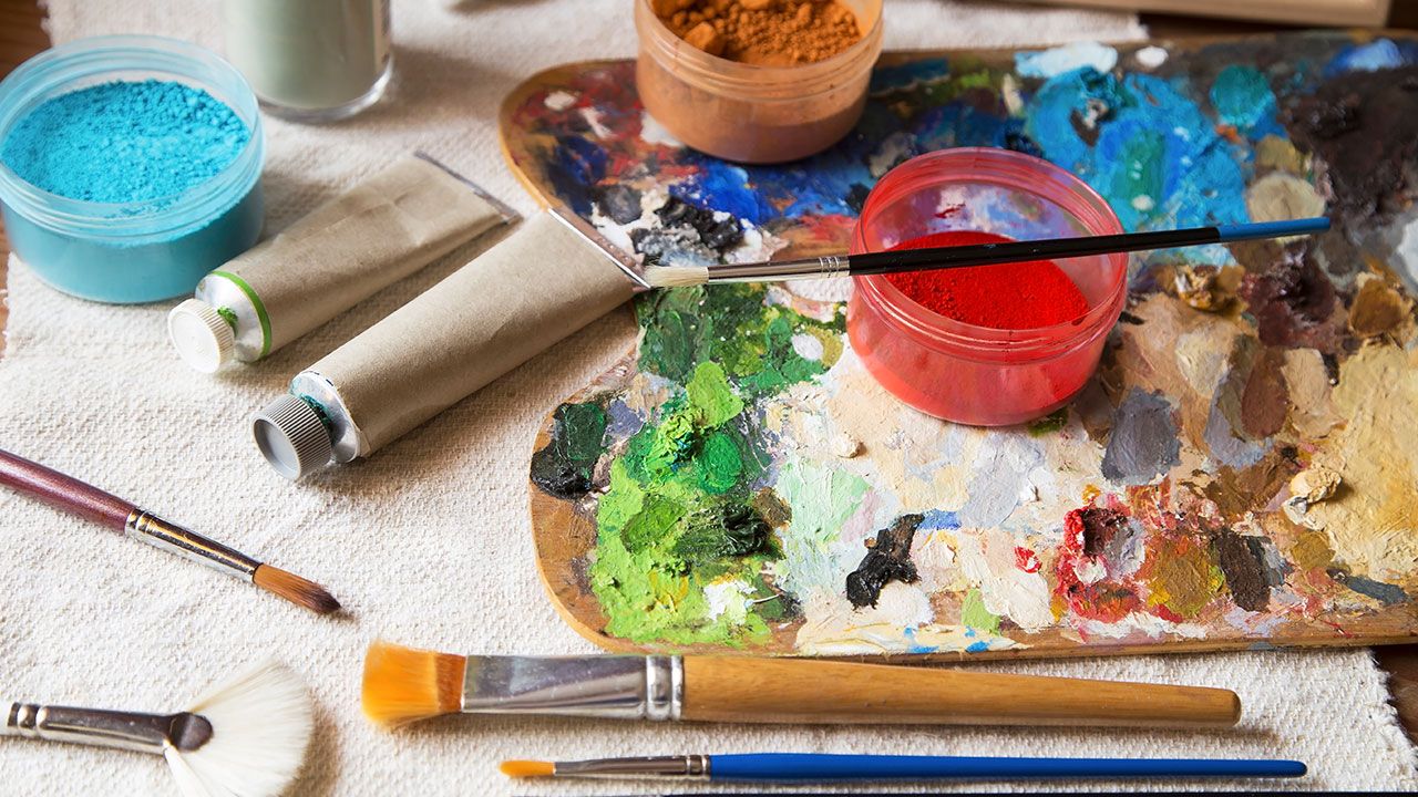 A painter's palette with colorful paints, brushes, and paint tubes on a textured cloth surface.