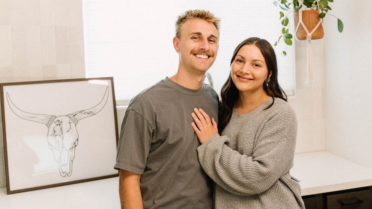 A smiling couple stands close together in a room, with a framed bull skull drawing and a hanging plant in the background.