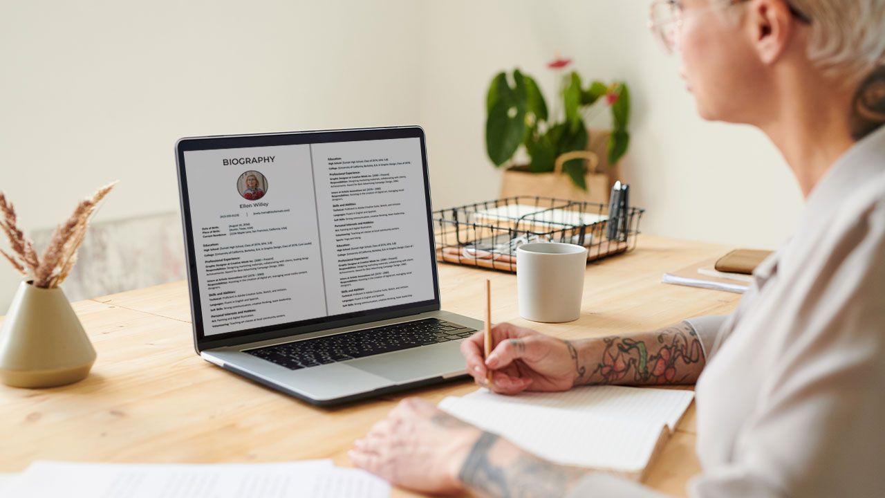 Person with tattoos writing notes at a wooden table, viewing a biography on a laptop. Nearby are a mug, papers, and a plant.