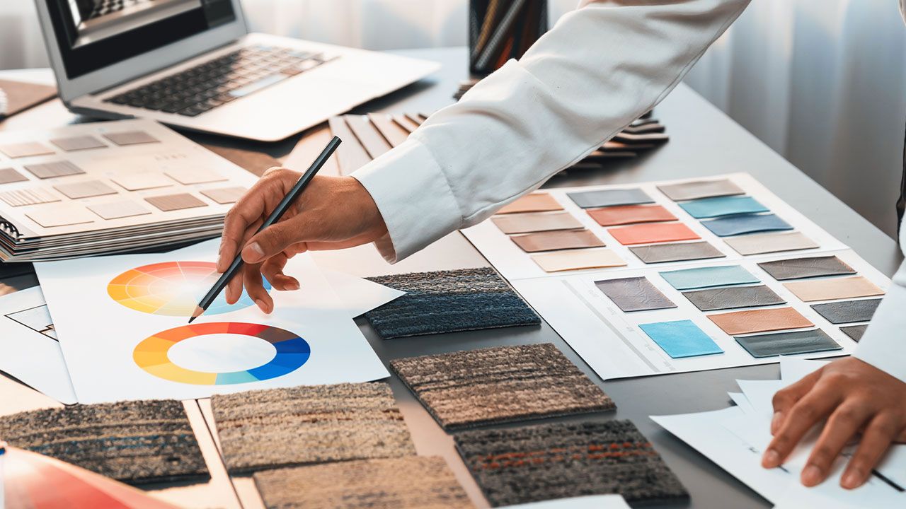 Person examining color palettes and fabric swatches on a table with a laptop, focusing on design planning and material selection.