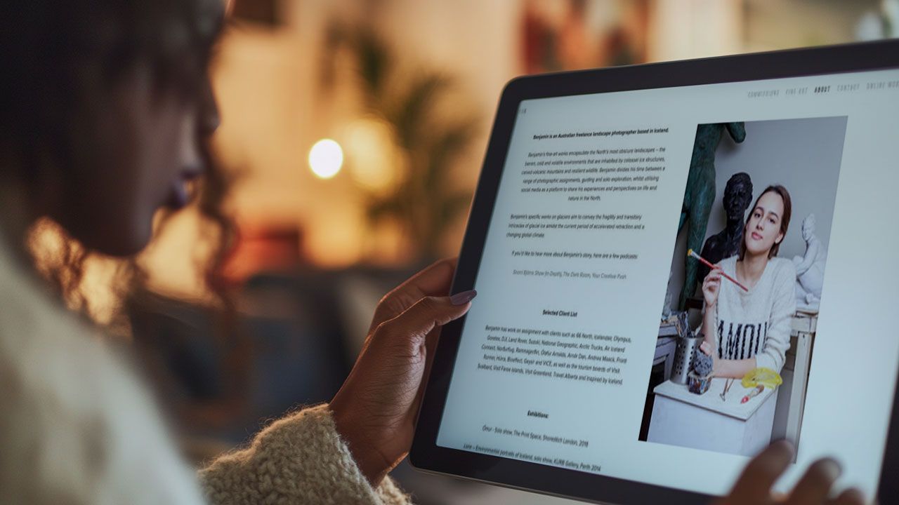 Person reading an article on a tablet, featuring an image of a woman with a paintbrush beside a sculpture. Blurred background.