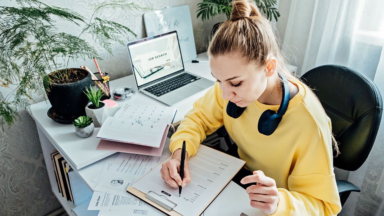 Woman in yellow sweater writing on a clipboard at a desk with a laptop, headphones, and plants.