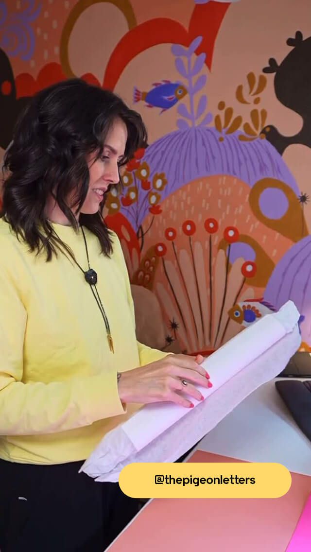 Woman in yellow shirt holding paper, standing in front of a colorful, abstract mural with floral and geometric patterns.