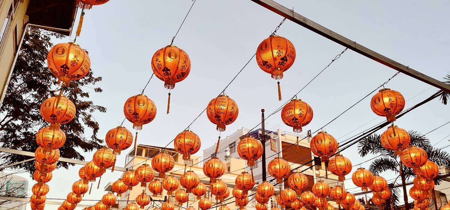 Numerous orange lanterns hanging in rows against a clear sky, with trees and buildings in the background.