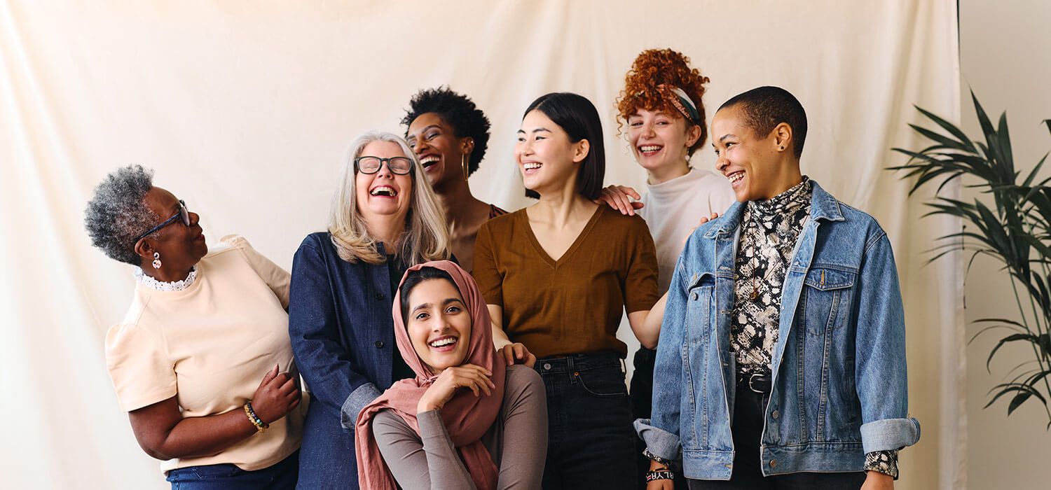 A diverse group of seven women smiling and laughing together in front of a neutral backdrop, with a plant on the side.