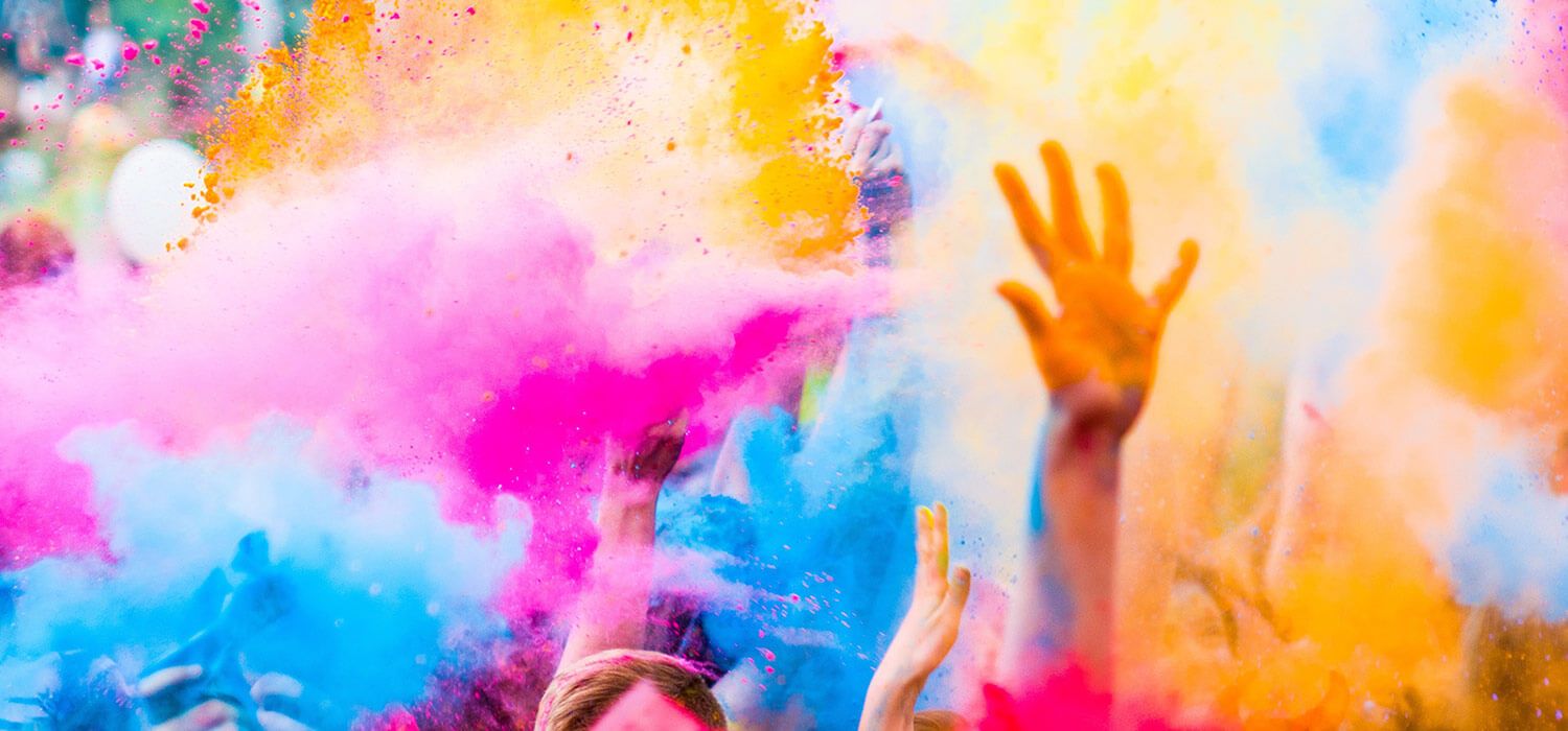 Vibrant festival scene with people throwing colorful powders in the air, creating a cloud of pink, yellow, and blue hues against a blurred background.