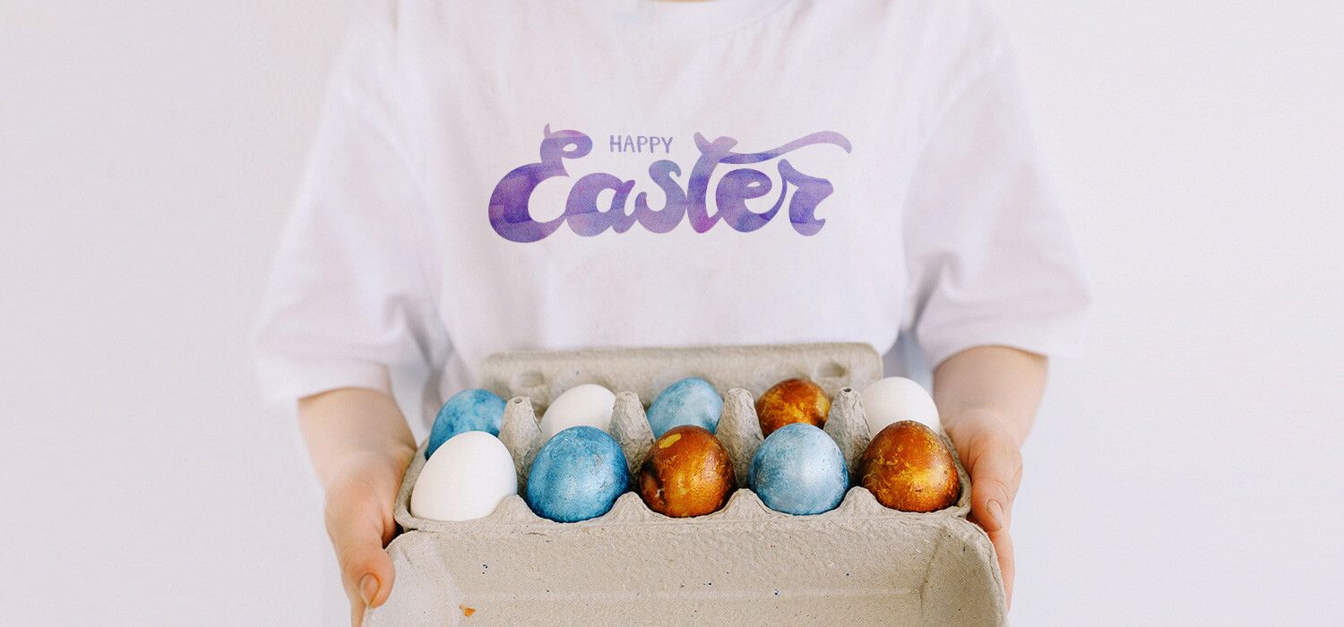 Person holding a carton of colorful Easter eggs, wearing a white shirt with "Happy Easter" written on it.