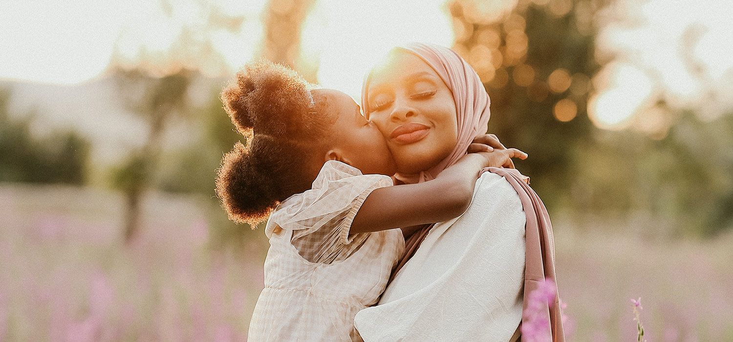 A woman in a hijab embraces a young child who kisses her cheek in a sunlit field, surrounded by soft-focus greenery and pink flowers.