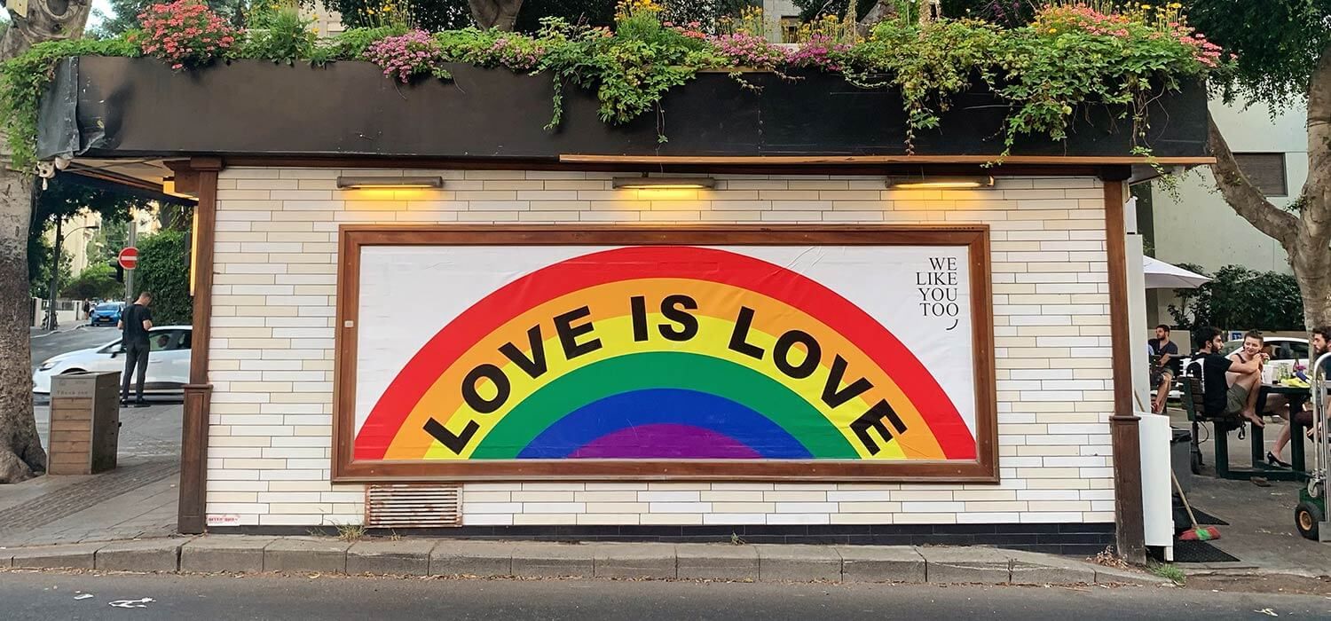 A mural with a rainbow and the words "LOVE IS LOVE" on a white brick wall, surrounded by greenery and outdoor seating.