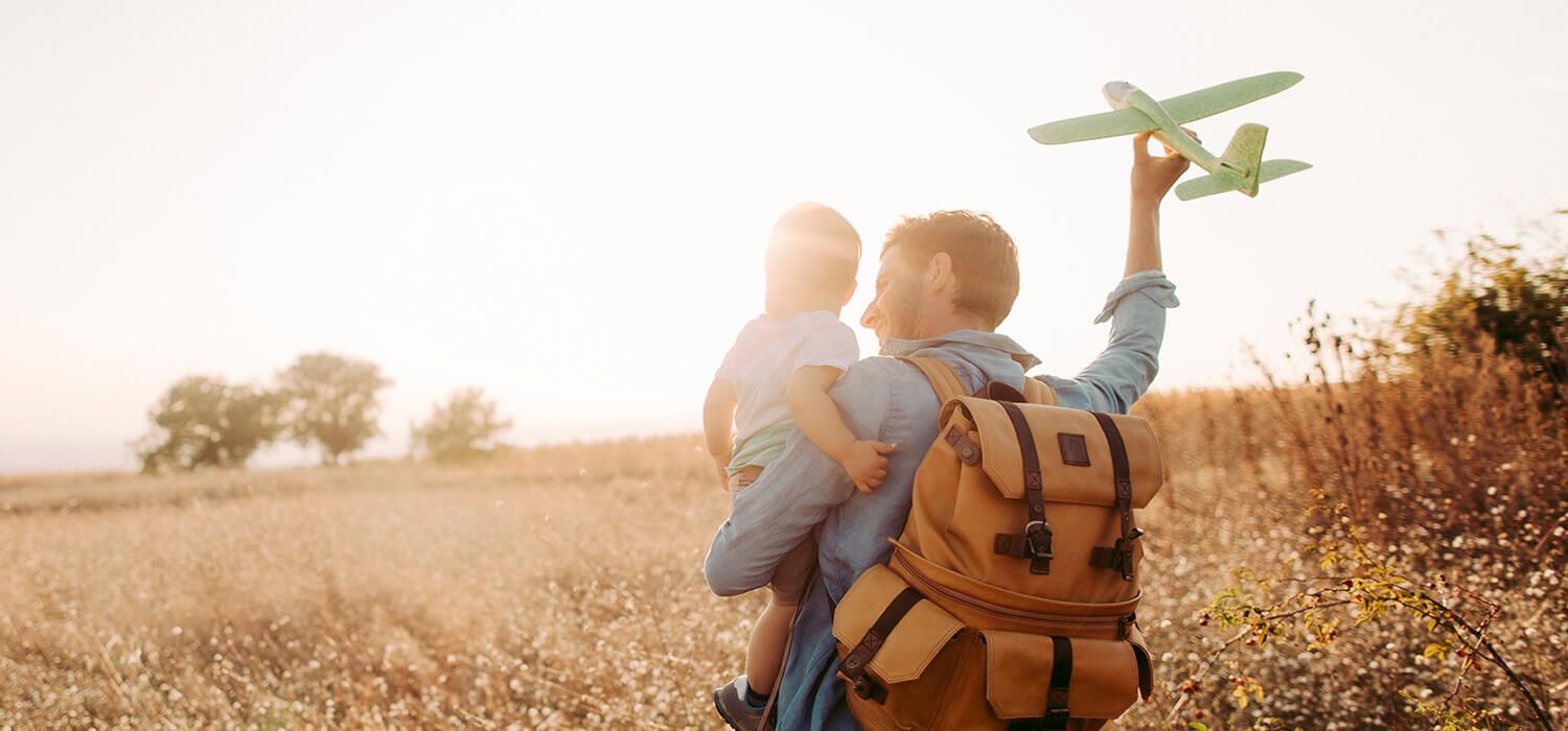 A person carries a child and holds a toy airplane in a sunlit field, wearing a backpack. Trees are visible in the background.