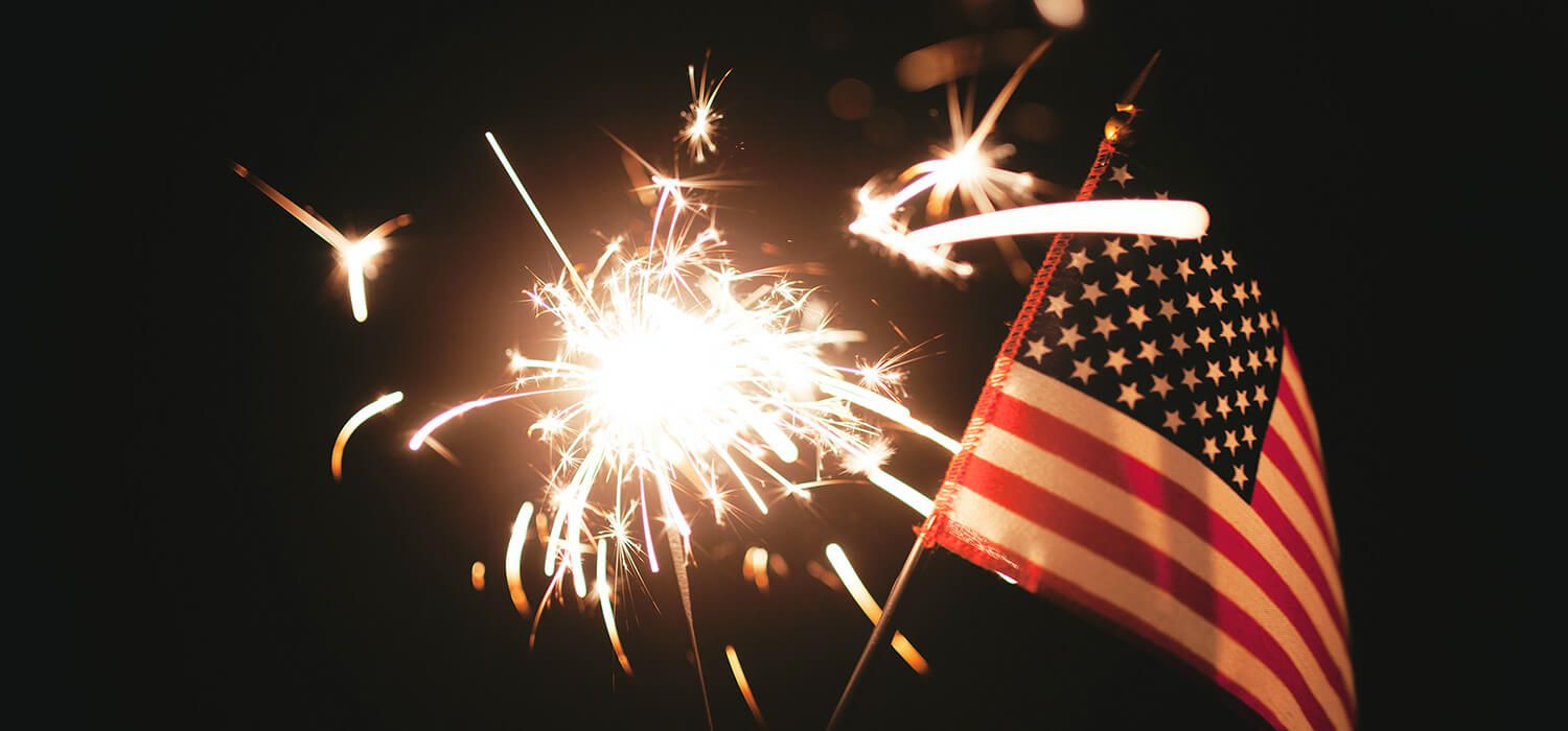 A lit sparkler illuminates a small American flag against a dark background.