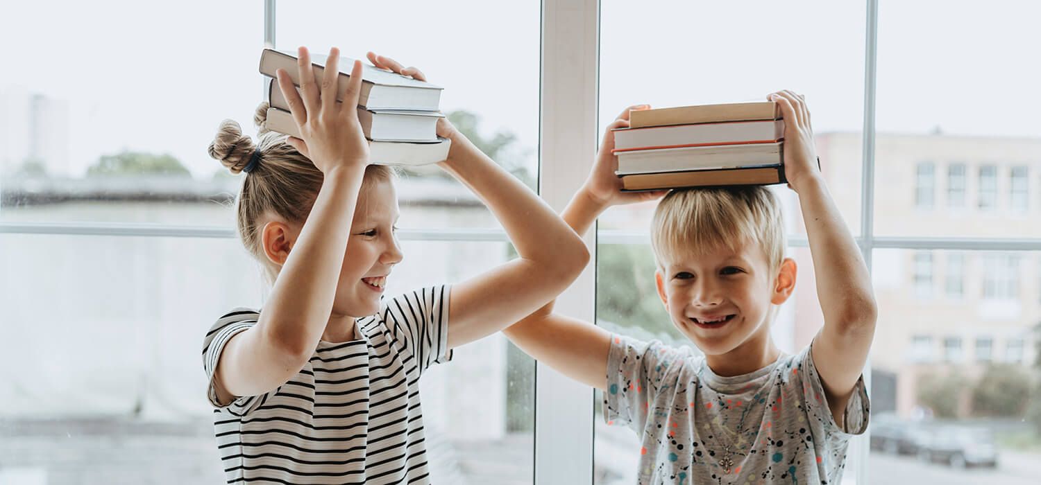 Two children smiling, balancing stacks of books on their heads, in front of a large window.
