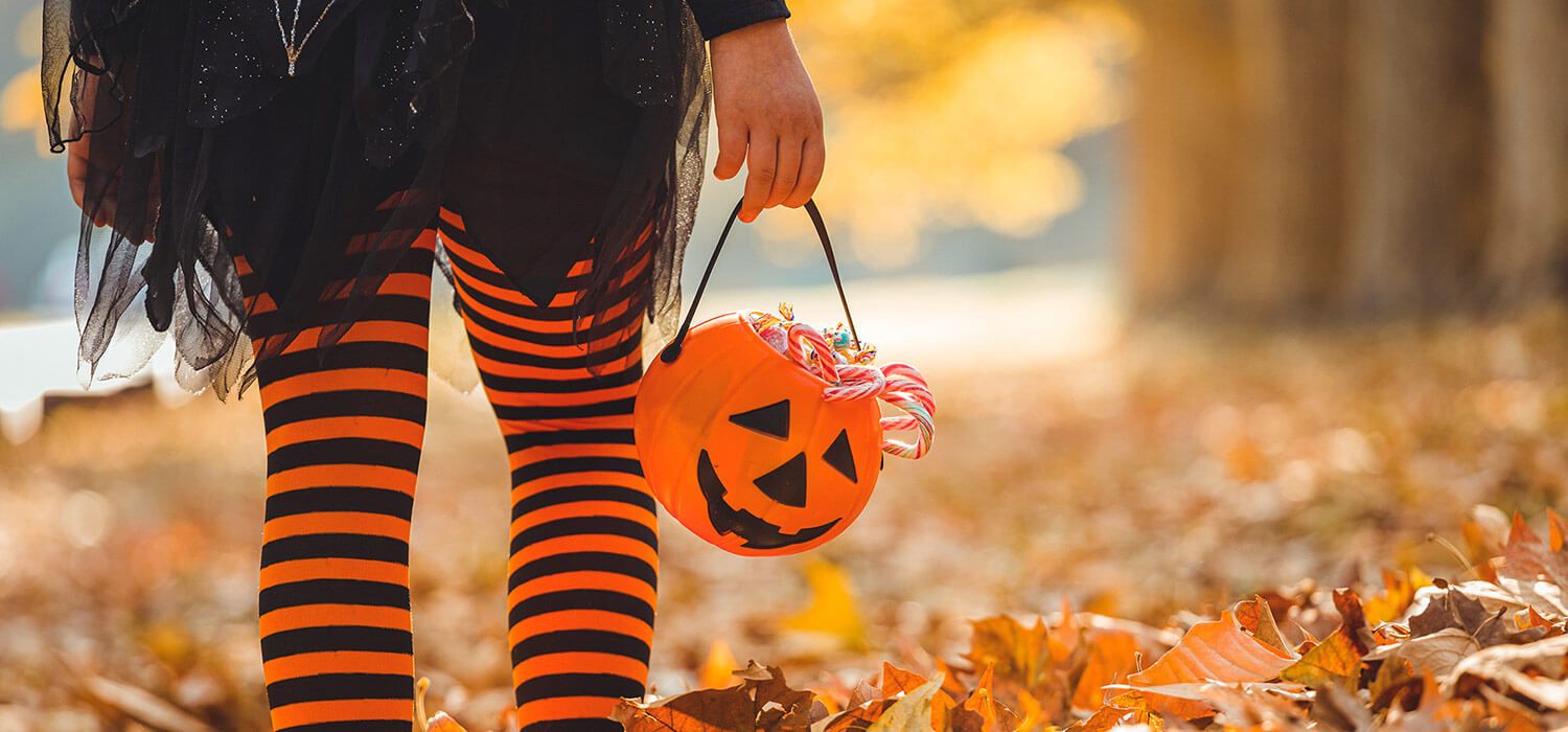 Child in striped leggings holds a pumpkin-shaped candy bucket, standing on fallen leaves in a park during autumn.