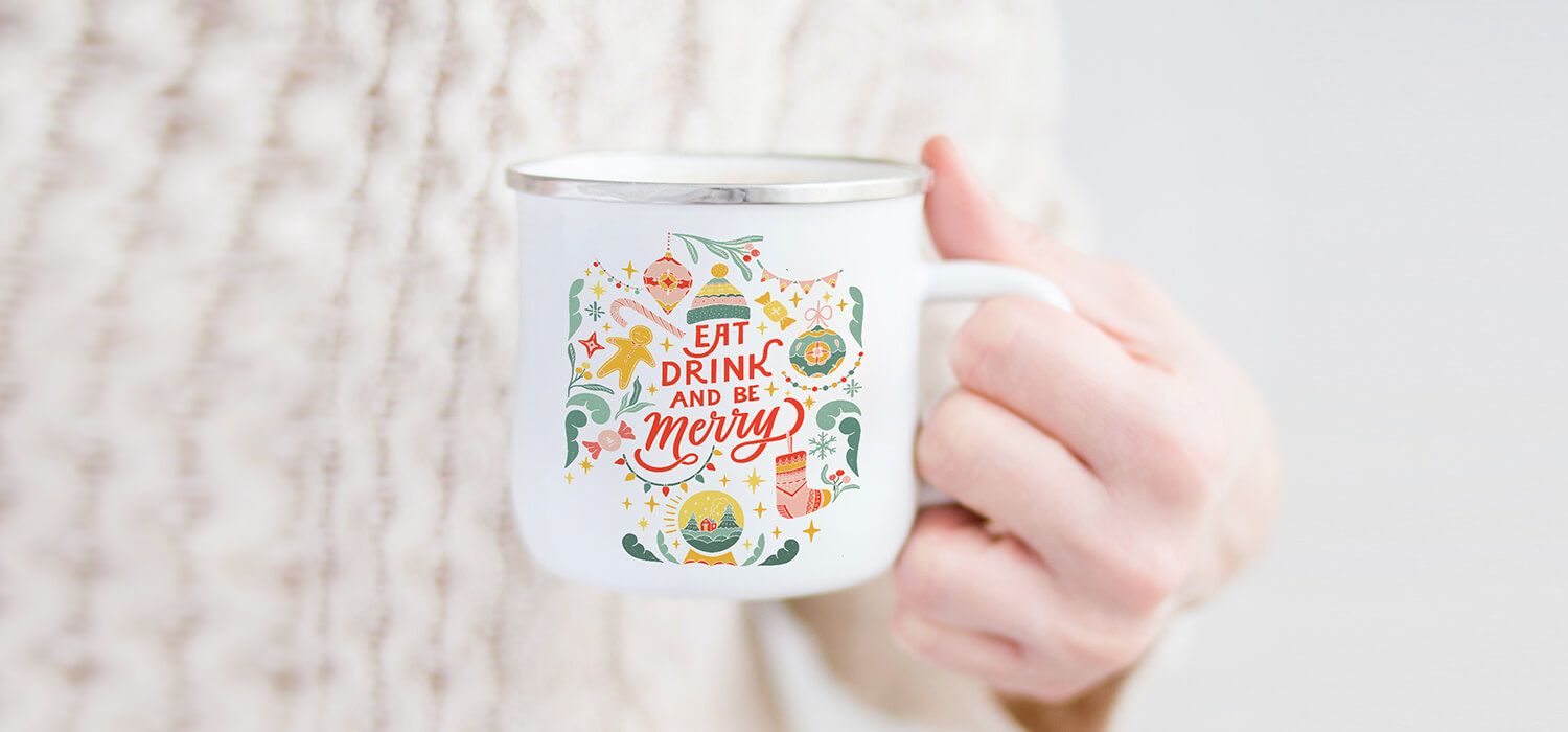 Hand holding a white mug with festive illustrations and the text "Eat, Drink and Be Merry" against a cozy, knitted sweater background.