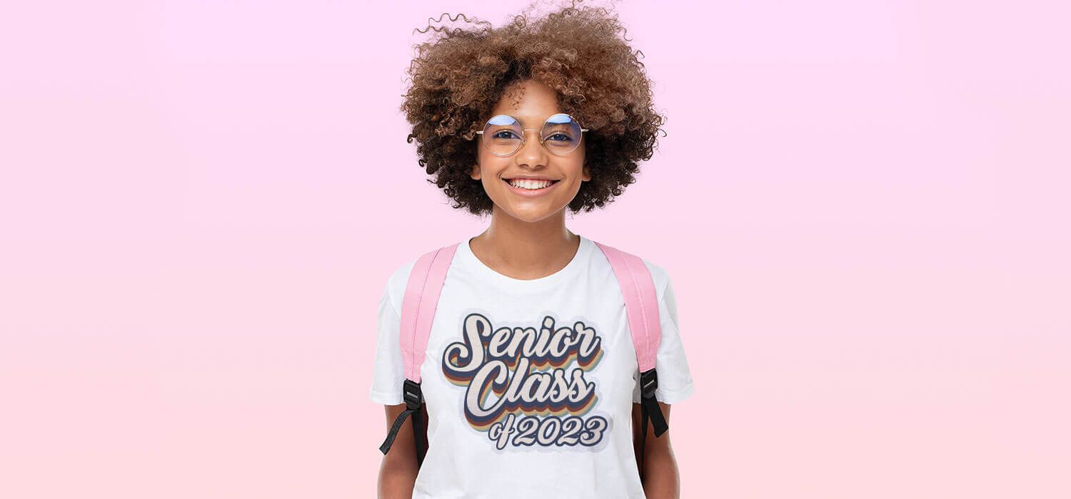 Smiling person with curly hair and glasses, wearing a "Senior Class of 2026" t-shirt and a pink backpack, against a pink background.