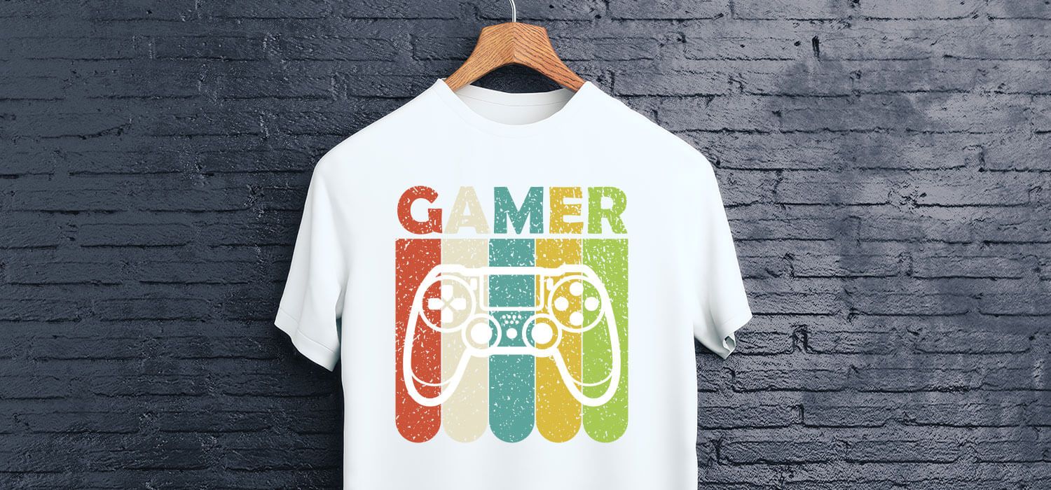 White t-shirt with a colorful "GAMER" design and a game controller graphic, hanging on a wooden hanger against a dark brick wall.