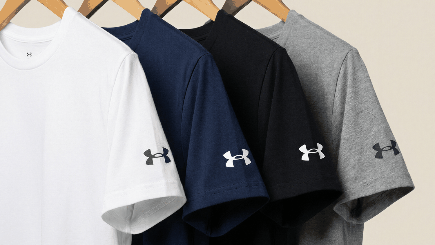 Four Under Armour crew‑neck t-shirts—white, navy, black, gray—hanging on wooden hangers with logos on the sleeves.